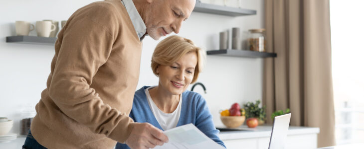 Middle aged couple checking finance account together. Wife and husband fill tax form, holding current expense document in hands, sitting on kitchen, paying utility bill online