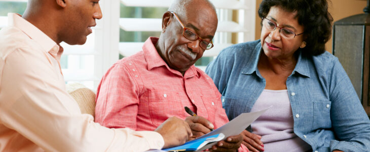 Financial Advisor Talking To Senior Couple At Home Signing Documents Sitting On Sofa