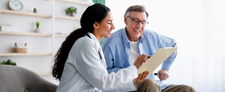 Young doctor asking senior impaired male patient in wheelchair to sign insurance policy at home. Handicapped elderly man putting his signature under surgery consent form, reading medical document