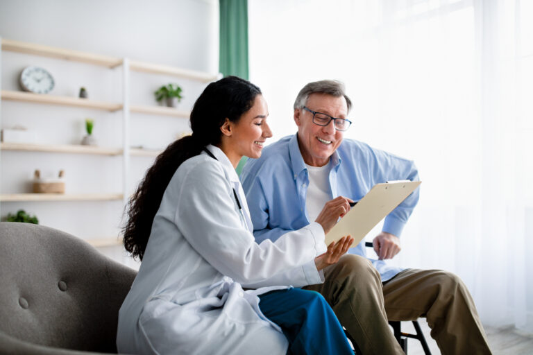 Young doctor asking senior impaired male patient in wheelchair to sign insurance policy at home. Handicapped elderly man putting his signature under surgery consent form, reading medical document