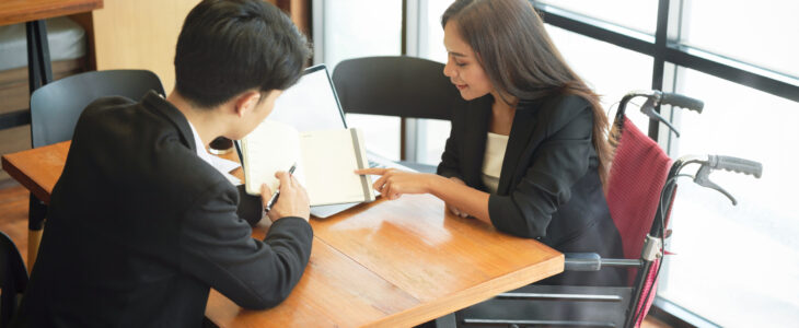 Disabled female business employee in wheelchair brainstorms with coworkers, listen colleague explain work process on notebook in office