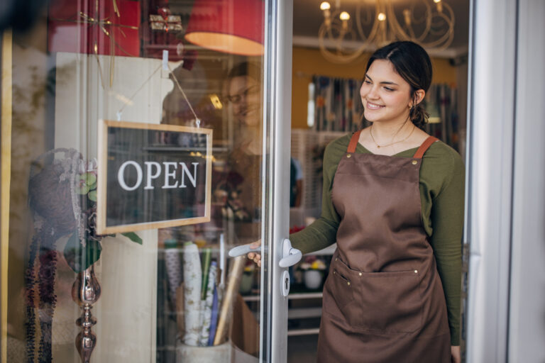 A young florist dressed in a brown apron and green shirt turns the 'CLOSED' sign to 'OPEN' on the door of a flower shop, smiling warmly, and ready to start the day.