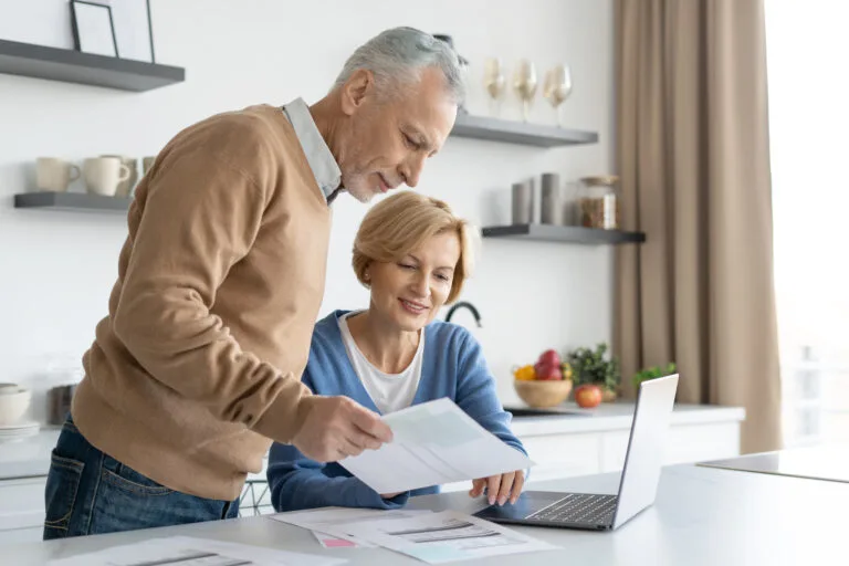 Middle aged couple checking finance account together. Wife and husband fill tax form, holding current expense document in hands, sitting on kitchen, paying utility bill online