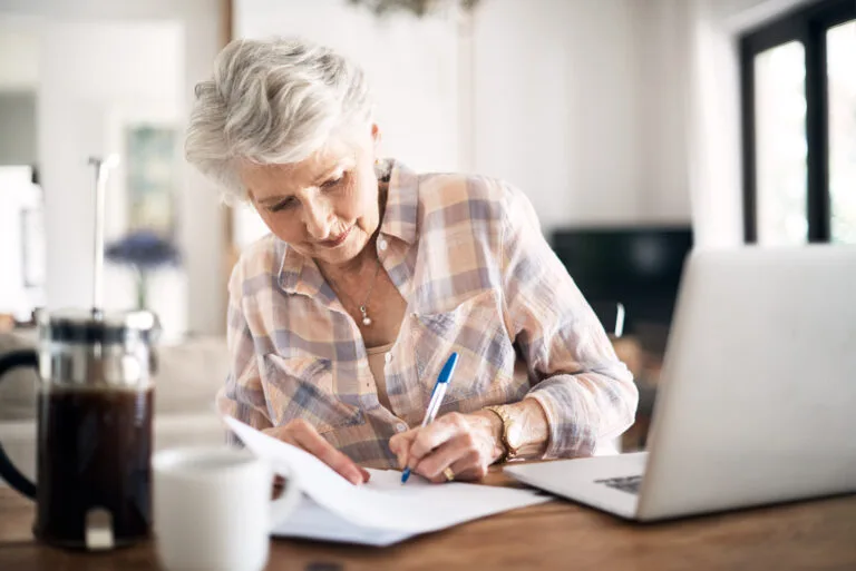 Shot of a happy senior woman flliing in paperwork while using her laptop at home