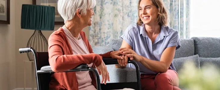 Mature woman comforting senior mom sitting on wheelchair at nursing home. Cheerful woman talking to old disabled mother in wheelchair at elder care centre. Loving caregiver taking care of elderly woman at home.