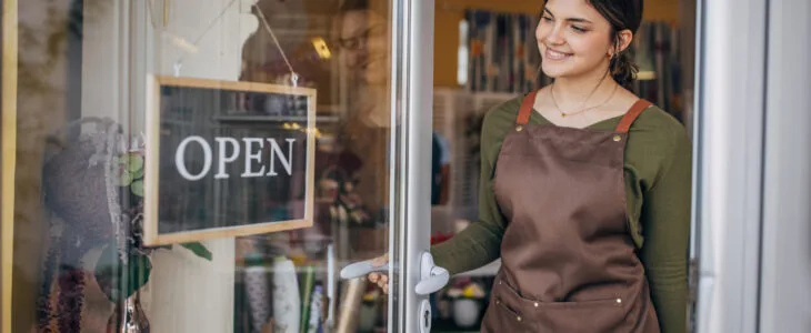 A young florist dressed in a brown apron and green shirt turns the 'CLOSED' sign to 'OPEN' on the door of a flower shop, smiling warmly, and ready to start the day.